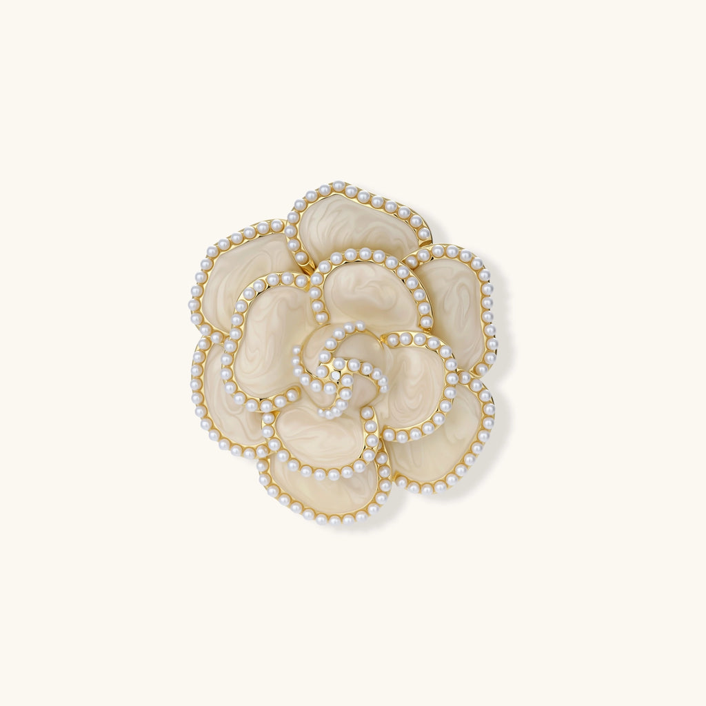 Cream enamel camellia flower brooch with gold-plated petal edges lined with tiny white round beads, on a plain cream background.