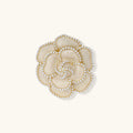 Cream enamel camellia flower brooch with gold-plated petal edges lined with tiny white round beads, on a plain cream background.